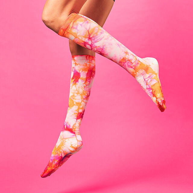 Fuchsia Flame pink and orange tie dye compression socks on woman’s legs, suspended in the air on a pink background